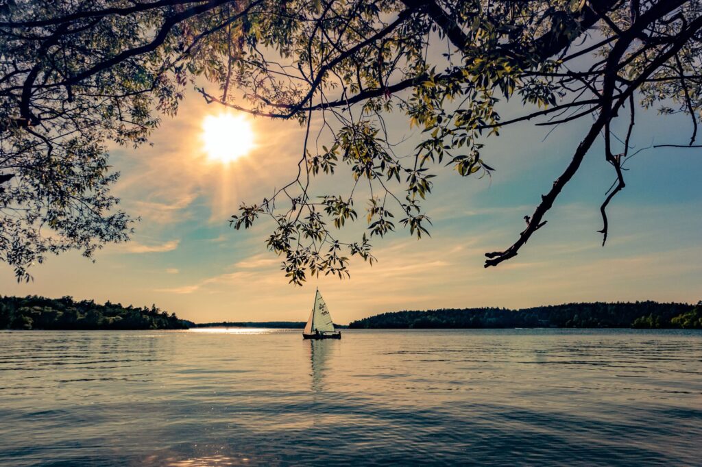 A wide landscape photograph of a small sailboat floating on calm water under a bright, glowing sun