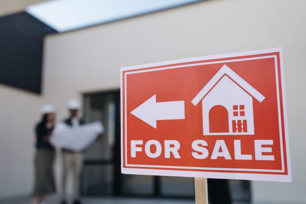A close-up, sharp-focus shot of a red and white "FOR SALE" sign with a white arrow and house icon pointing to the left